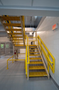 Yellow grated metal staircase and railings inside an industrial facility, with an illuminated exit sign on the wall to the right.