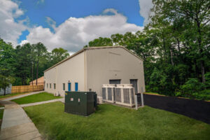 Industrial metal building with beige siding and large rooftop HVAC units in a grassy area, surrounded by trees and a blue sky with clouds.