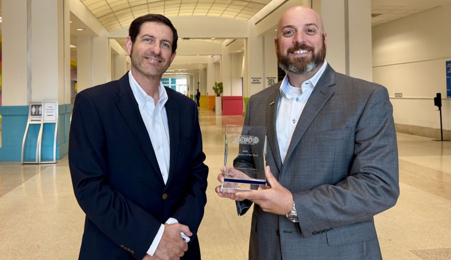 Two men in business attire pose in a bright hallway, holding a glass award and smiling at the camera.
