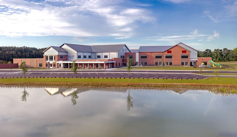 Wide exterior of a modern elementary school with brick and white siding, phased wings, and a large parking lot under a bright blue sky with clouds.