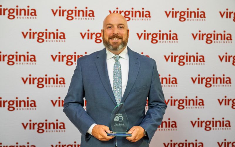 Man in a blue suit smiles while holding a glass award, with a Virginia Business backdrop behind him.