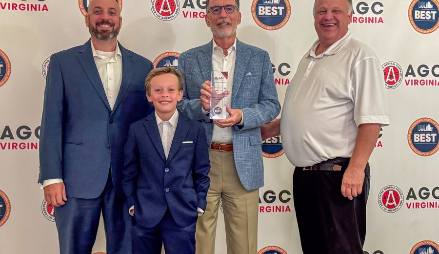 Four men in suits pose for a group photo at an AGC Virginia event, holding a glass trophy with a backdrop of AGC Virginia and BEST logos.