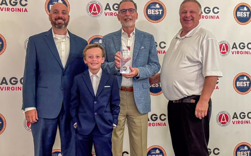 Four men in suits pose for a group photo at an AGC Virginia event, holding a glass trophy with a backdrop of AGC Virginia and BEST logos.