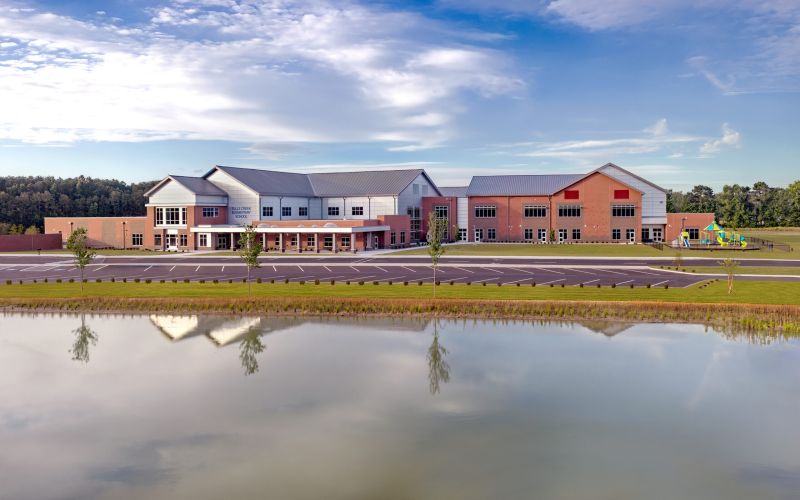 Wide exterior of a modern elementary school with brick and white siding, phased wings, and a large parking lot under a bright blue sky with clouds.