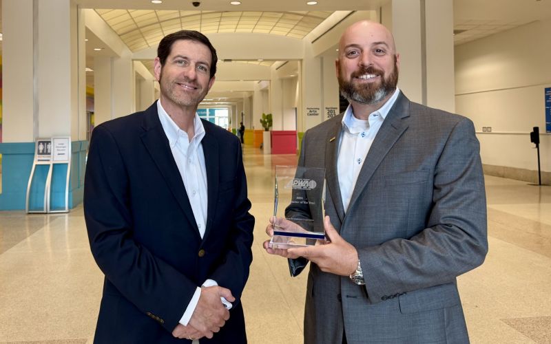 Two men in business attire pose in a bright hallway, holding a glass award and smiling at the camera.