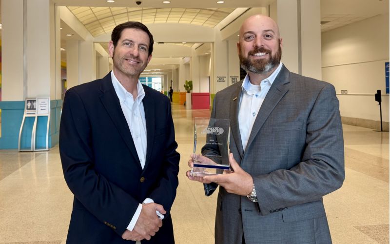Two men in business attire pose in a bright hallway, holding a glass award and smiling at the camera.